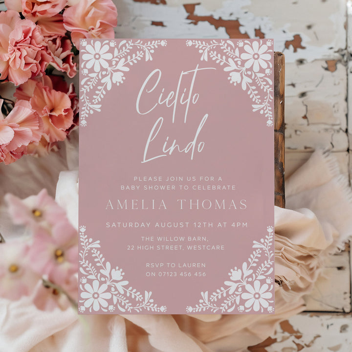 Soft pink fiesta-themed baby shower card with Cielito Lindo calligraphy and botanical accents, styled on woven background.