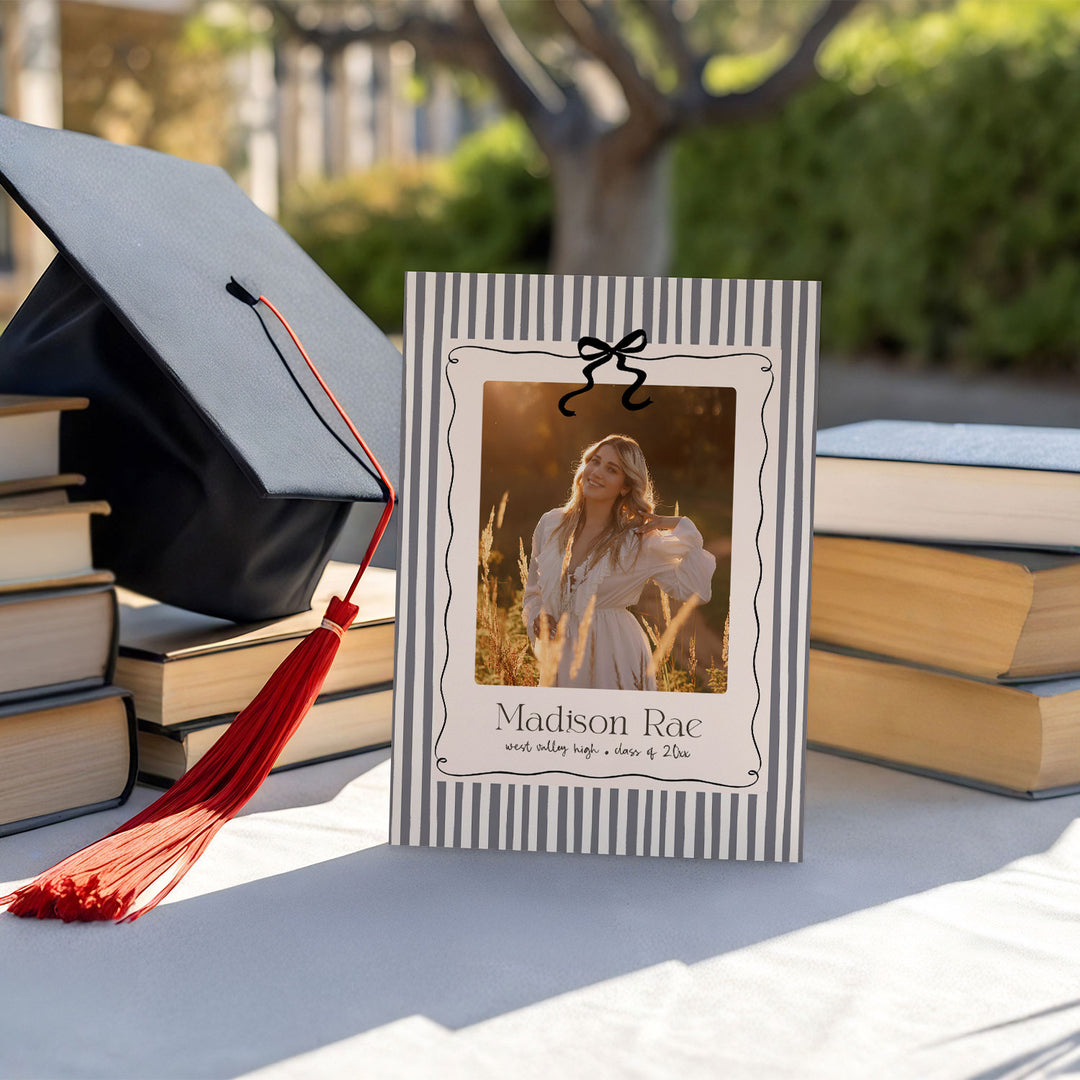 Graduation cap, books, and a photo frame with a personalized message on a table outdoors.
