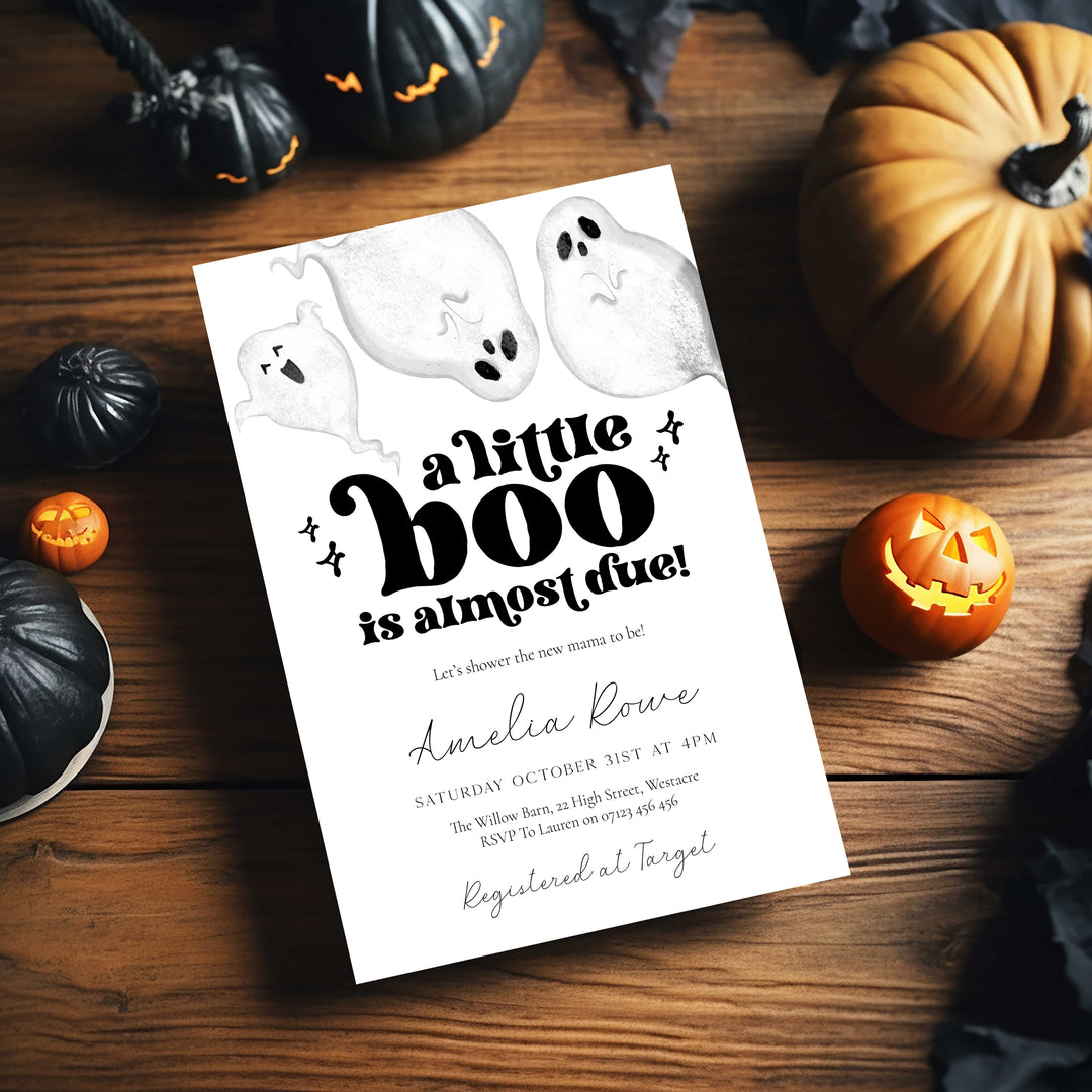 Halloween-themed baby shower invitation with ghosts and pumpkins on a wooden table.