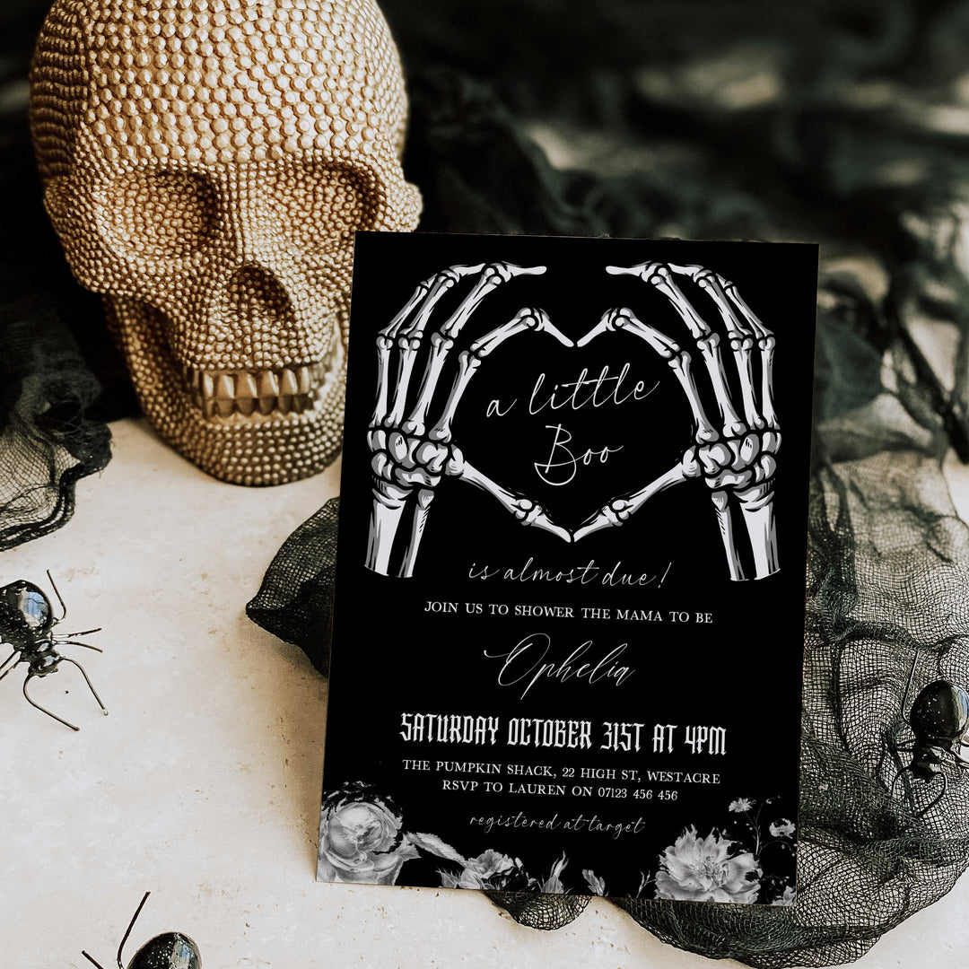 Halloween-themed baby shower invitation with skeleton hands forming a heart on a black background, placed on a decorative surface with a skull and spider.