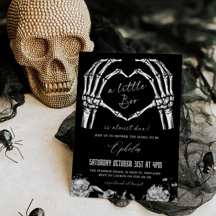 Halloween-themed baby shower invitation with skeleton hands forming a heart on a black background, placed on a decorative surface with a skull and spider.
